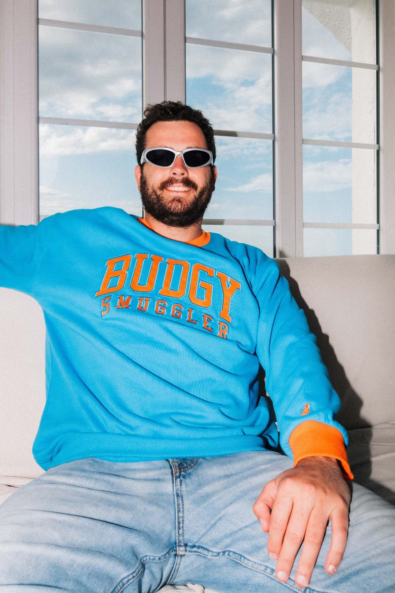 Man and Women wearing blue crewneck jumper with orange writing on front, in a lounge room 