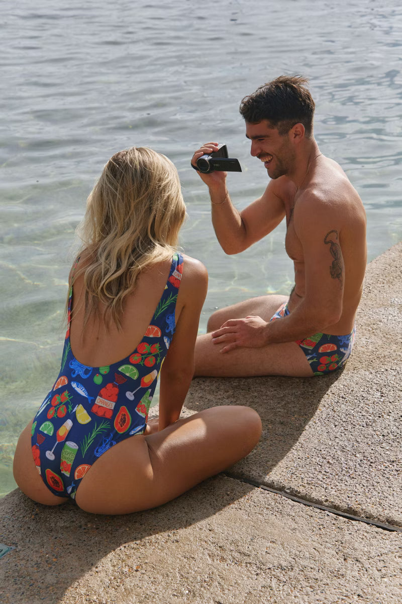 Girl wearing Blue women's one piece with fruit pattern by the water with male wearing the same in swim briefs 